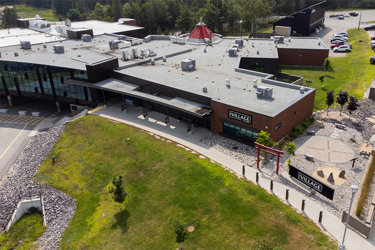 Aerial view of The Village, showcasing modern buildings with flat roofs, green spaces, parking lots, and a landscaped area featuring a red torii gate and rock garden.